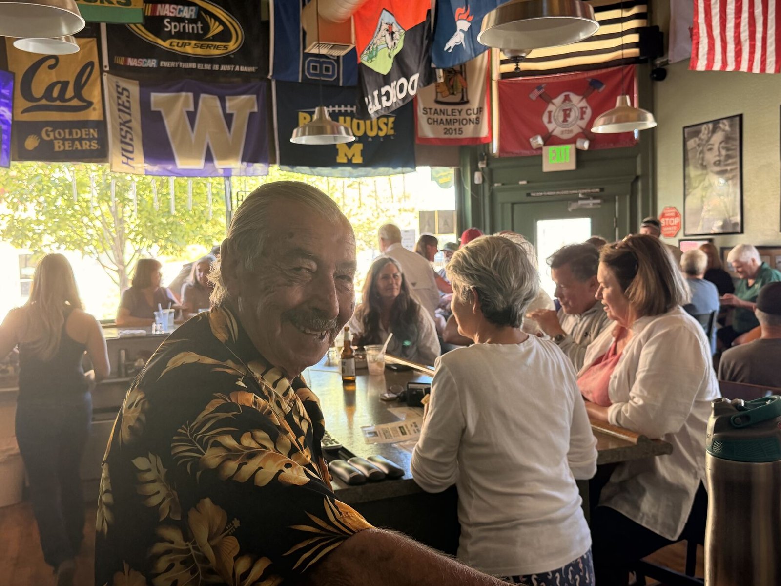 A smiling regular in a Hawaiian shirt turns around on his stool while the bar fills up